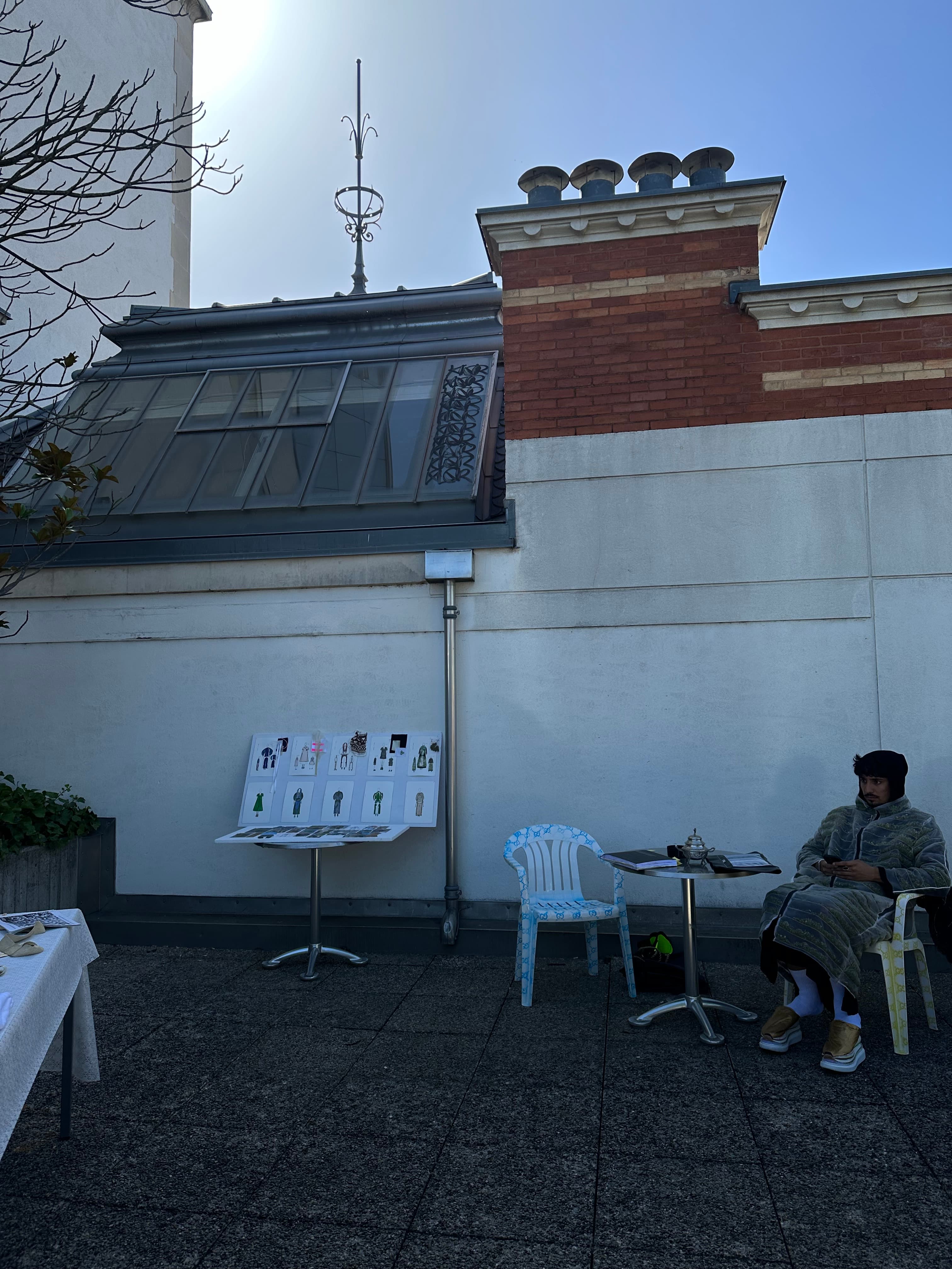 Rooftop setup with illustrated look boards on a stand, a Moroccan teapot on a café table and a model waiting in a quilted striped coat