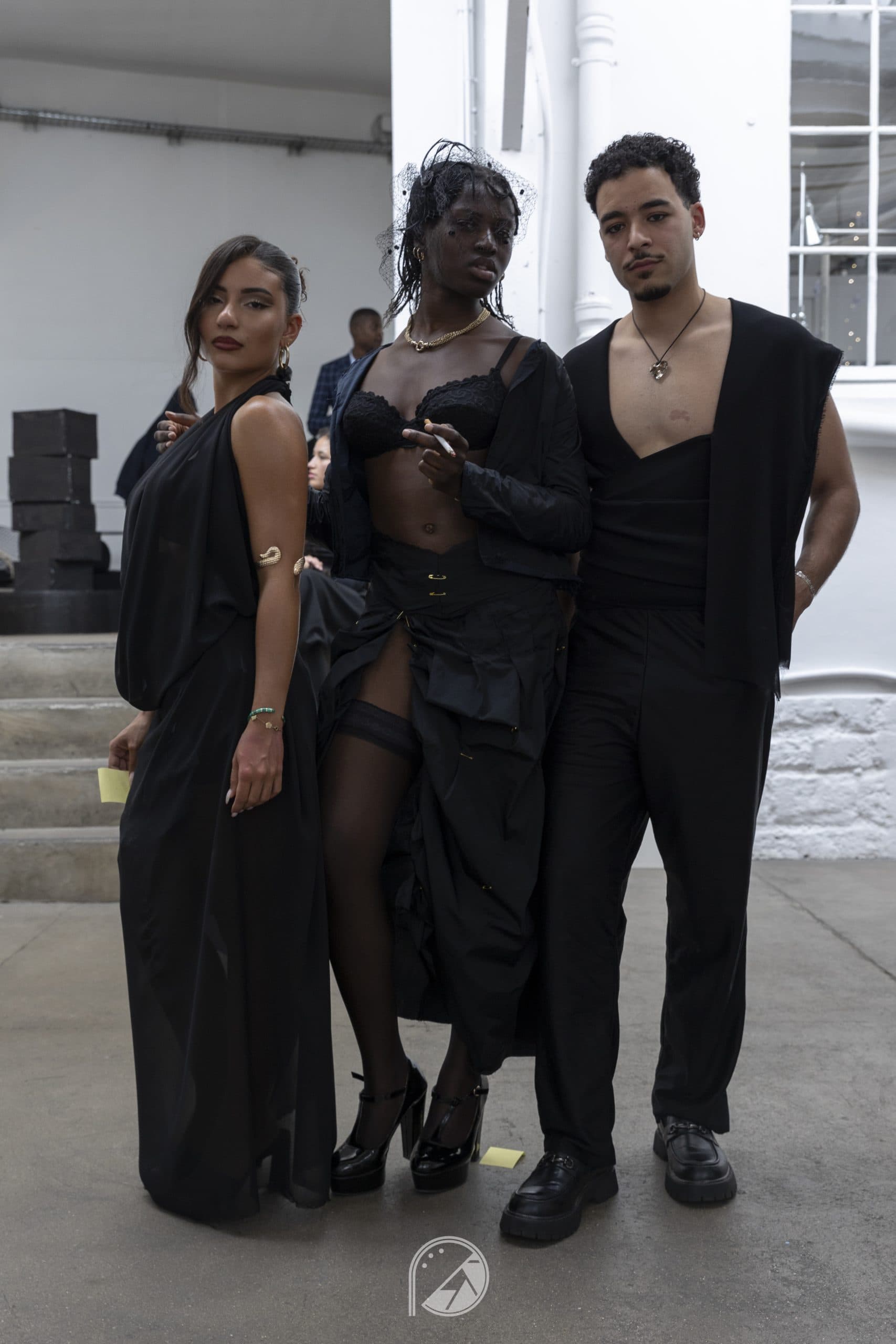 Three models in all-black looks with lace, draping and safety pins, posing backstage