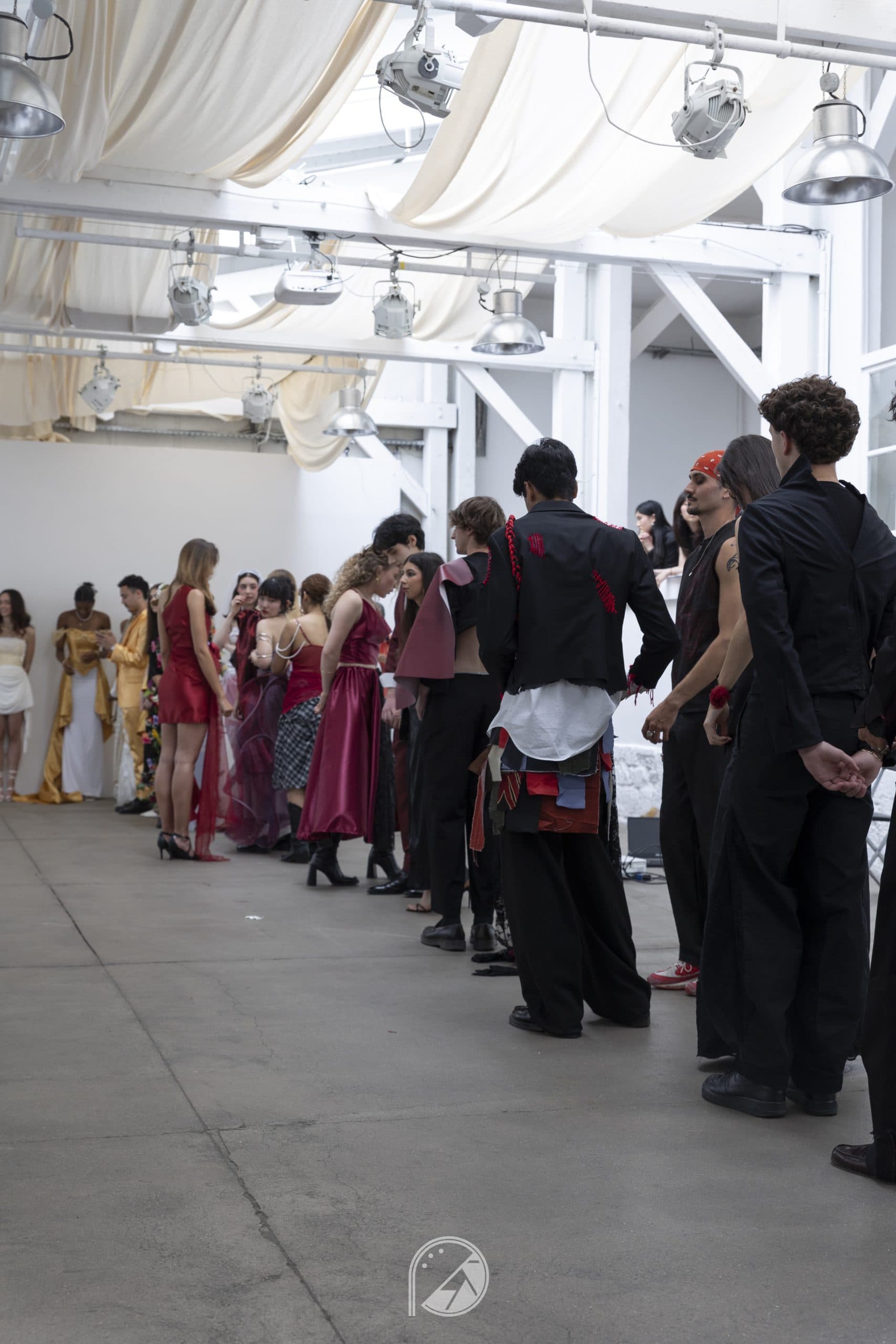 Models in red, gold and black looks standing in a long line-up inside the white industrial space of Le QG