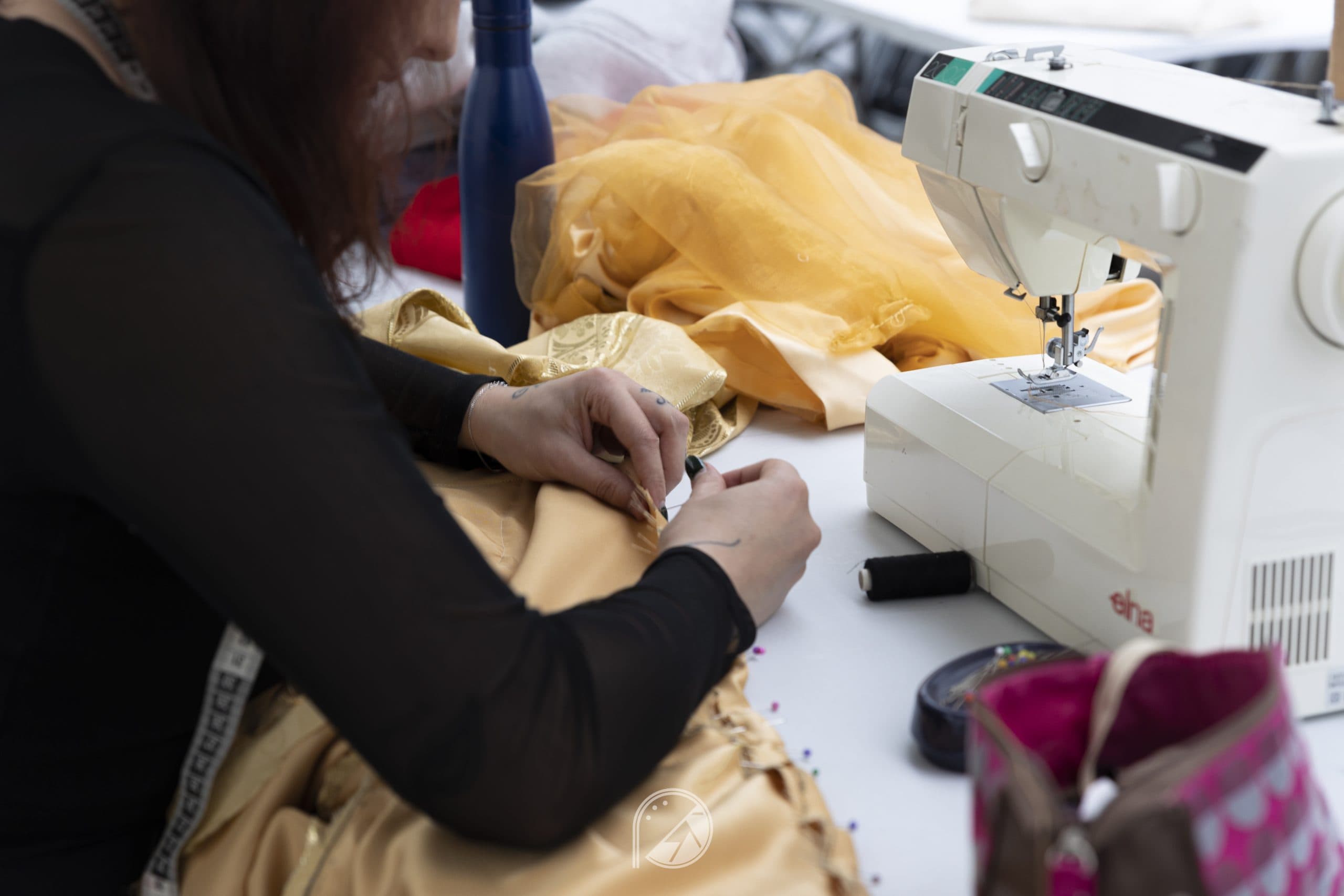 Hands pinning golden satin beside a sewing machine during last-minute alterations
