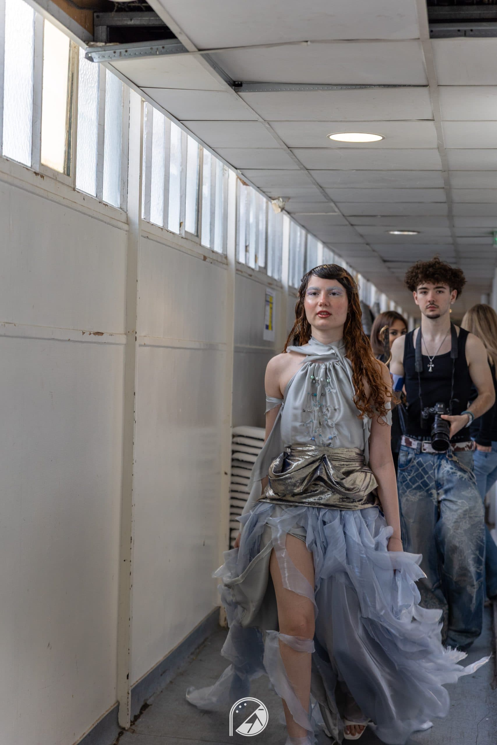 Model walking down the backstage corridor in the full silver and lilac gown, a photographer behind her